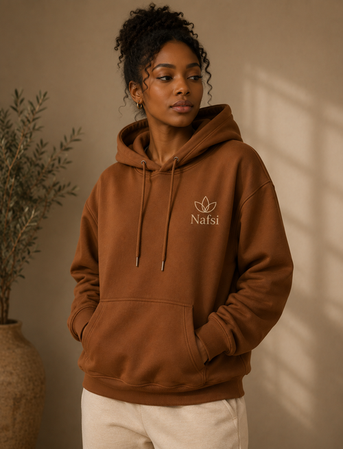 A woman wearing a brown pullover hoodie with a white embroidered logo that reads 'Nafsi' and a leaf emblem on the left chest, posed in a warm, neutral-toned indoor setting.